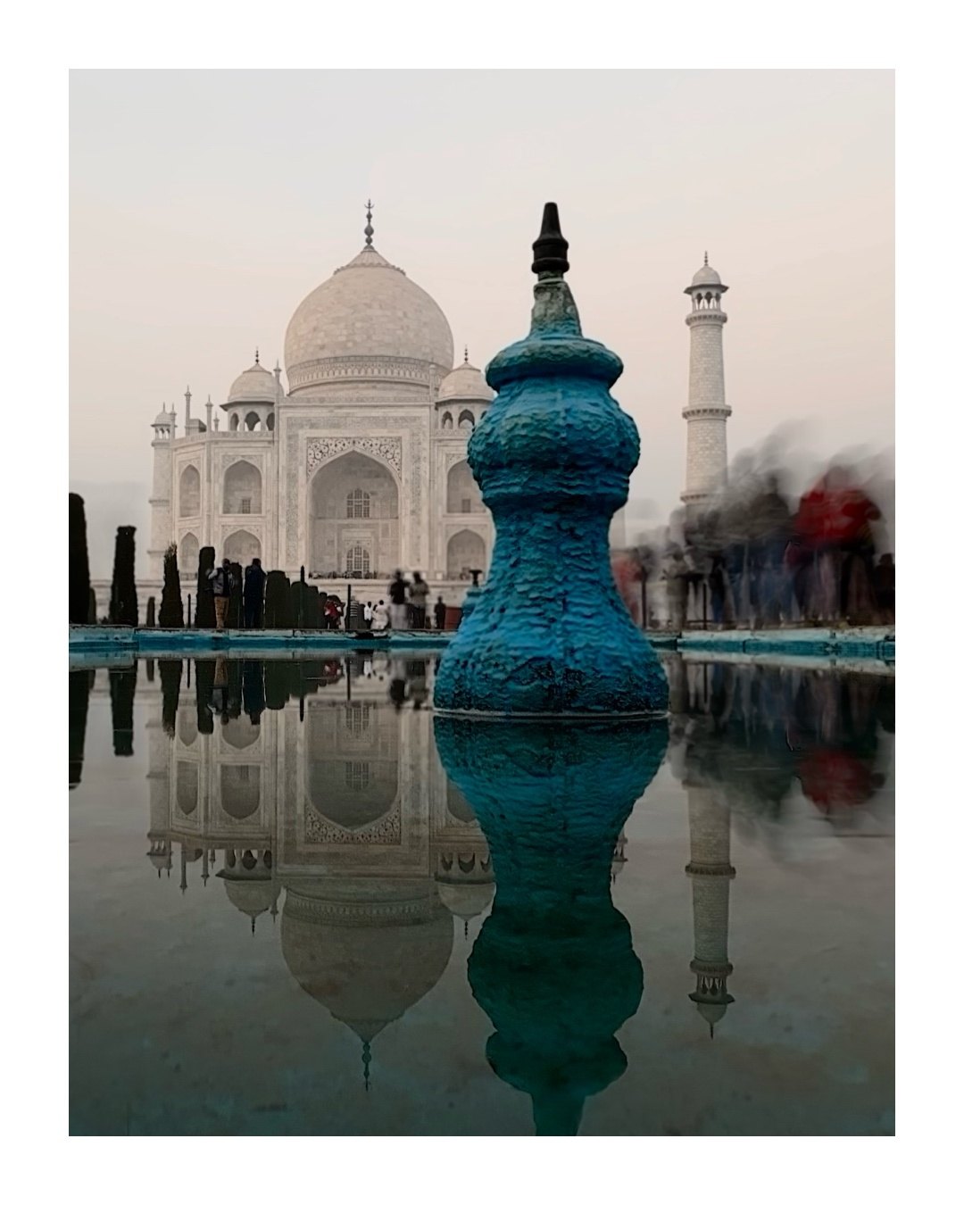 The Taj Mahal at dusk behind a blue-painted finial, with its reflection in the pool and blurred figures at the right