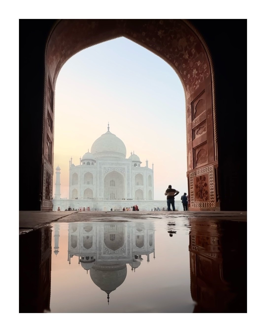 The Taj Mahal seen through a large carved arch at sunrise, with its reflection in a puddle
