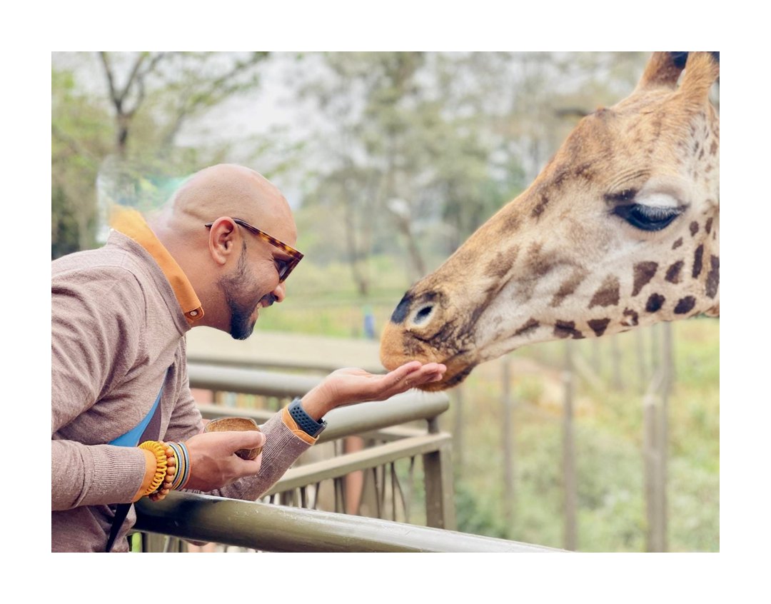 Photo of a man in sunglasses leaning over a railing and smiling as a giraffe eats from his open hand