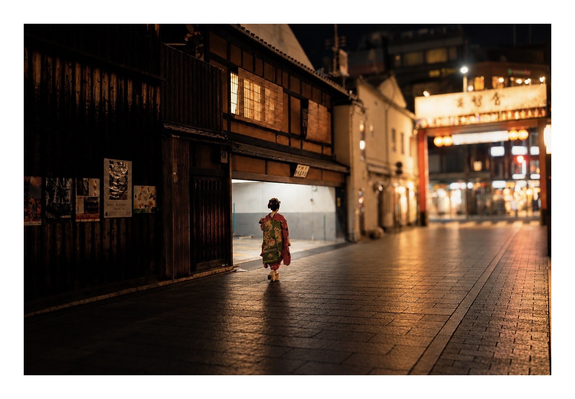 A maiko in a red kimono walks away down a wet stone street in Kyoto at night, toward a glowing orange gate